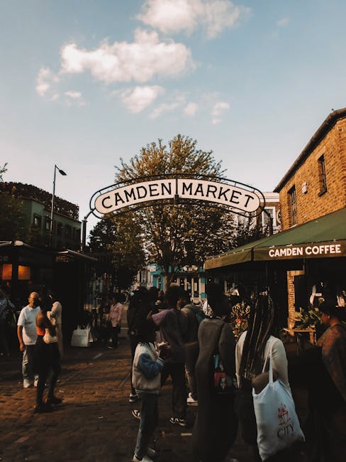 Image of a busy urban street scene in Camden, NW1, showing a variety of buildings including shops and residential structures with decorative facades and large windows. The street features marked lanes, crosswalks, and traffic signage, with pedestrians walking along sidewalks and a motorcycle parked at the curb. The sky is partly cloudy, casting natural light over the scene. The overall atmosphere is lively and well-maintained, reflecting typical city activity. This image is related to the service offered by Carpet Cleaners Camden, emphasizing the importance of surface cleanliness and maintenance in both domestic and commercial environments to ensure hygiene and presentation, aligned with the focus of NW1 same day carpet cleaning availability in Camden.